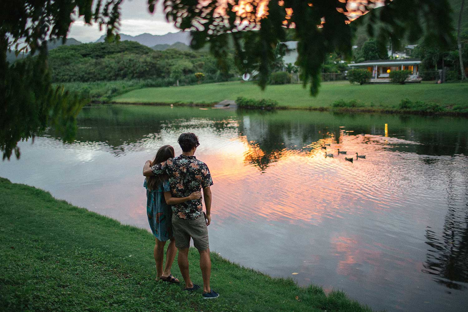 man and woman with arms around another in front of pond at sunset