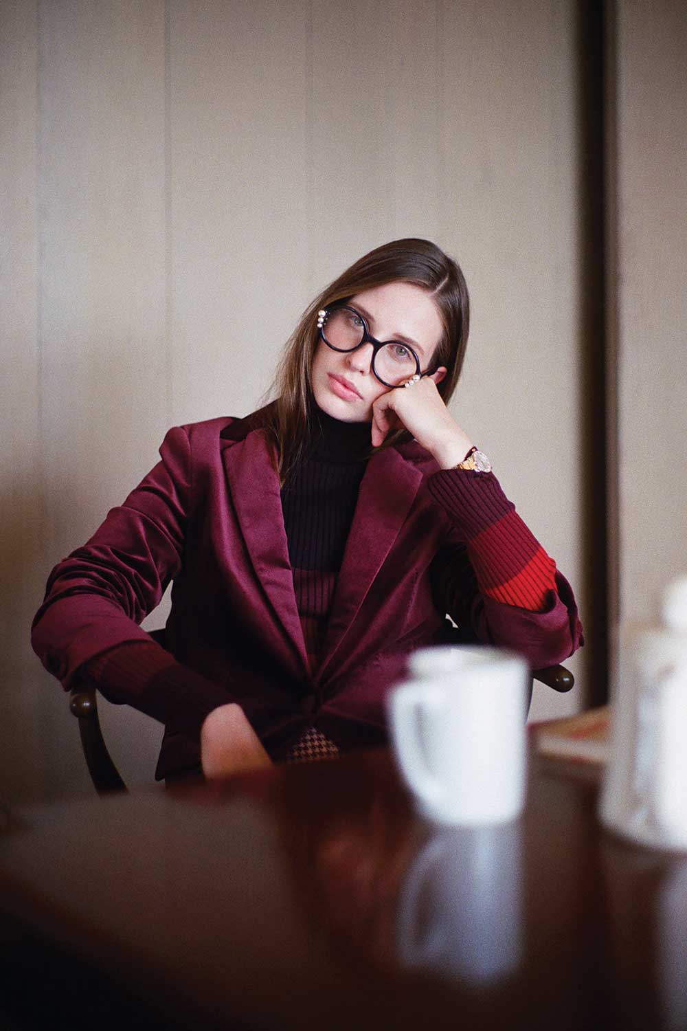 Woman in maroon velvet blazer with round glasses on, coffee in foreground on table