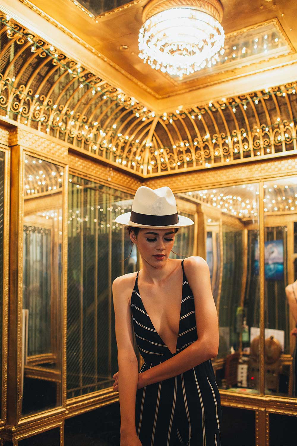 Woman in black striped dress and white hat standing in gold mirrored elevator
